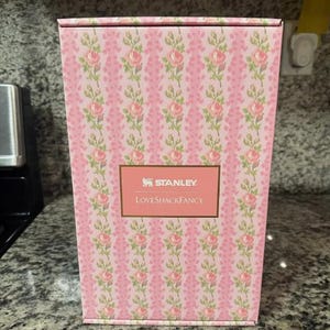 May include: A pink rectangular box with a floral pattern of pink roses and green leaves. The box has a gold label with the text "STANLEY" and "LOVESHACKFANCY".