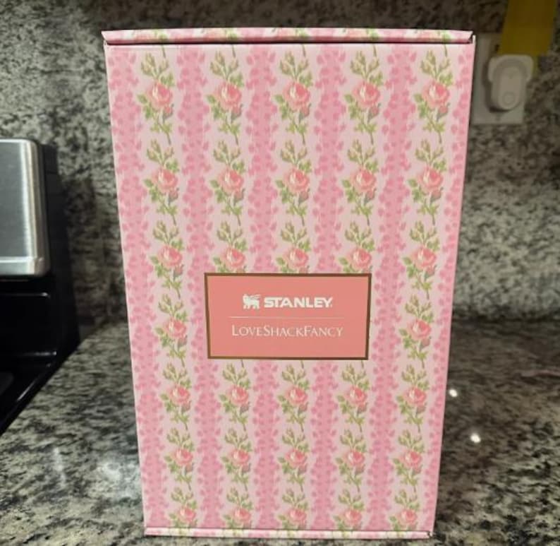 May include: A pink rectangular box with a floral pattern of pink roses and green leaves. The box has a gold label with the text "STANLEY" and "LOVESHACKFANCY".