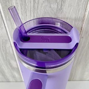 Puede incluir: Un vaso de color lavanda con tapa y pajita moradas. La tapa tiene un cierre deslizante y un asa. El vaso tiene una banda de acero inoxidable cerca de la parte superior y un logotipo en la tapa. La pajita est&aacute; en &aacute;ngulo.