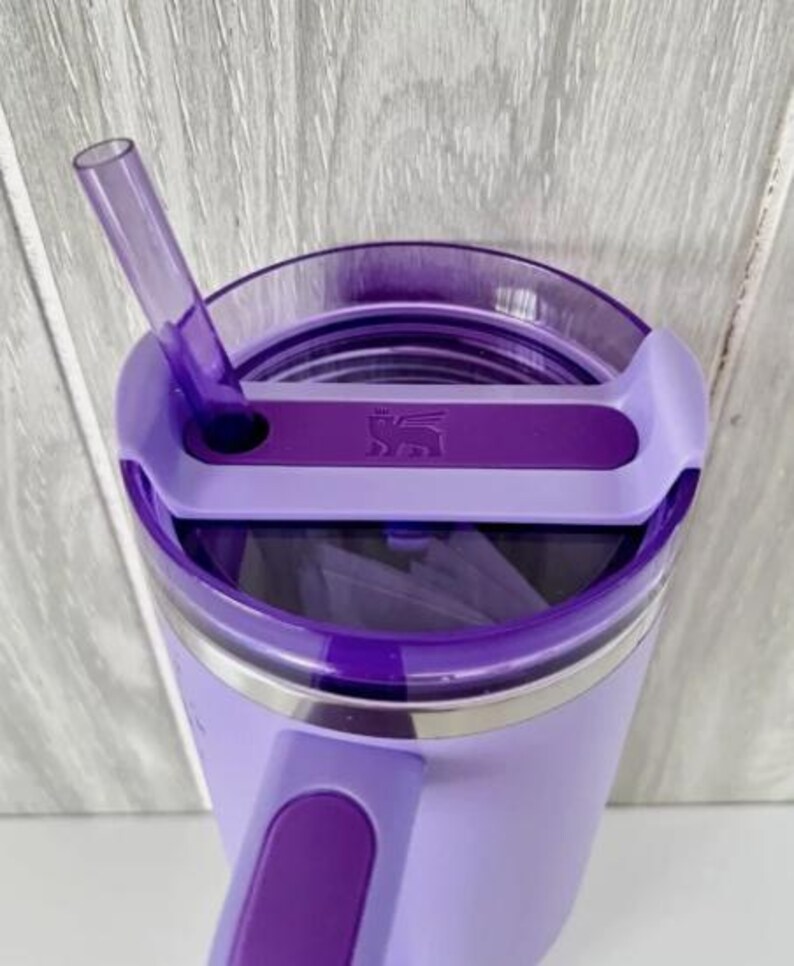 Puede incluir: Un vaso de color lavanda con tapa y pajita moradas. La tapa tiene un cierre deslizante y un asa. El vaso tiene una banda de acero inoxidable cerca de la parte superior y un logotipo en la tapa. La pajita est&aacute; en &aacute;ngulo.