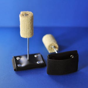 Op de afbeelding: Twee vuur-performance rekwisieten met een cilindrische beige lontkop bevestigd aan een dunne metalen staaf. De staaf is vastgezet op een zwarte rechthoekige basis met vier zilveren klinknagels, en elke basis heeft een zwarte elastische band.