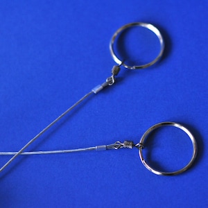 May include: Two silver-colored keychains with circular rings and thin wire attachments are displayed against a vibrant blue background. The keychains are positioned diagonally, creating an intersecting pattern.