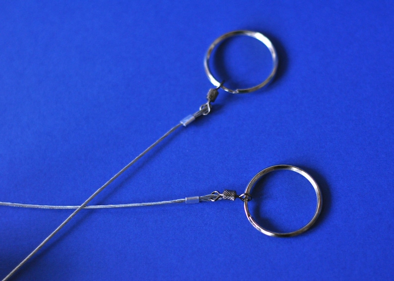 May include: Two silver-colored keychains with circular rings and thin wire attachments are displayed against a vibrant blue background. The keychains are positioned diagonally, creating an intersecting pattern.