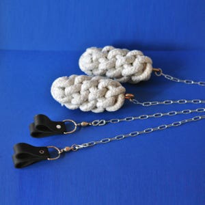 Peut inclure: Deux accessoires en corde grise tressée avec des chaînes argentées et des boucles en cuir noir. Les nœuds en corde sont reliés aux chaînes par des anneaux métalliques cuivrés. Le fond est bleu uni.