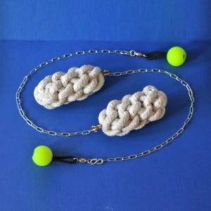 Pode incluir: Dois ornamentos de corda trançada esbranquiçada conectados por uma corrente prateada. Cada ornamento tem um pequeno fecho de latão. Duas bolas verde néon são presas à corrente com cordões pretos. O fundo é azul liso.