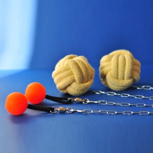 Peut inclure: Deux balles de jonglage orange vif avec des cordons noirs et des attaches à chaîne argentée au premier plan. Deux balles en corde beige sont à l'arrière-plan. Le tout sur fond bleu uni.