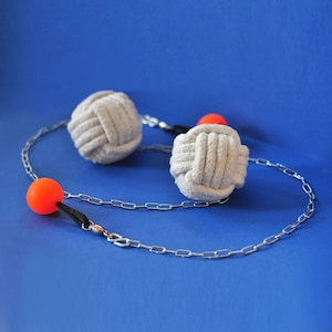 May include: A necklace featuring two gray, knotted rope balls and two orange spheres. The balls are connected by a silver chain and black cord, set against a blue background. The design is simple and modern.