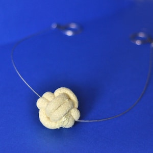 Könnte beinhalten: Eine cremefarbene Halskette mit Knotenanhänger aus Seil, einer dünnen Silberkette und zwei silbernen Schlaufen. Die Halskette befindet sich vor einem einfarbigen blauen Hintergrund, was einen auffälligen Kontrast erzeugt.