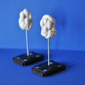 Peut inclure: Deux objets décoratifs avec un dessus gris noué, montés sur une fine tige métallique et une base rectangulaire noire. La base a une surface texturée noire et est fixée avec des rivets argentés. Le fond est bleu uni.