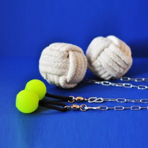 Op de afbeelding: Twee witte touwballen met zilveren kettingen en twee neon groene ballen met zwarte koorden. De objecten staan tegen een effen blauwe achtergrond. De touwballen zijn geknoopt en de kettingen zijn bevestigd met zilveren sluitingen.