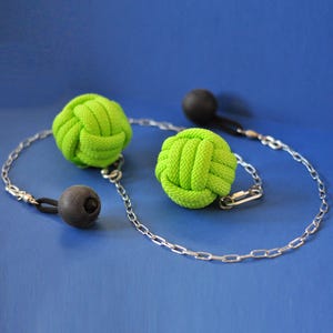 May include: A handcrafted necklace featuring two bright green knotted rope spheres and two black spheres. The spheres are connected by a silver chain. The necklace is set against a blue background.