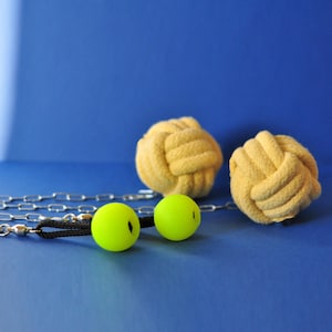 Puede incluir: Un conjunto de bolas de malabares amarillas y bolas de cuerda anudada sobre una superficie azul. Las bolas de malabares son de color verde brillante con detalles en negro. También se ven una cadena plateada y un cordón negro.