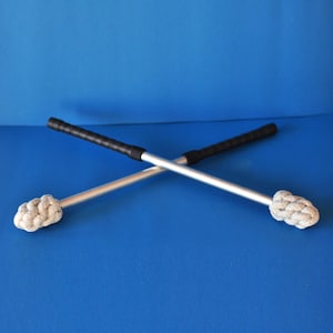 Op de afbeelding: Twee gekruiste drumstokken met zilveren schachten, zwarte handvatten en witte, geknoopte koppen. De stokken staan tegen een effen blauwe achtergrond, wat een eenvoudige, maar visueel opvallende compositie creëert.