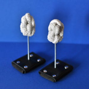 May include: Two fire poi heads featuring white cotton rope knots attached to thin metal rods. Each rod is secured to a black rectangular leather handle with four silver-coloured metal rivets. The construction includes a flexible black strap base.