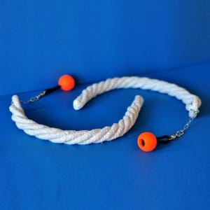 Op de afbeelding: Twee witte touwspeeltjes met oranje ballen en zilveren kettingen op een blauwe achtergrond. Het touw is dik en gedraaid en de oranje ballen zijn klein en rond. De kettingen verbinden het touw met de oranje ballen.