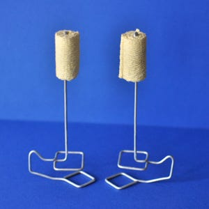 以下が含まれることがあります： ベージュの円筒形の芯頭と銀色の金属スタンドが付いた2つの火口。スタンドは、湾曲したデザインの長方形のベースを備えています。背景は無地の青色です。