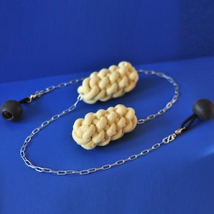 Op de afbeelding: Twee beige, gevlochten objecten verbonden door een zilveren ketting, elk met een zwarte bol aan het uiteinde. De objecten staan tegen een effen blauwe achtergrond. De ketting is gemaakt van kleine, verbonden metalen stukjes.