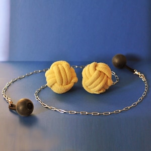 Peut inclure: Deux boules jaunes nouées en corde, reliées par une chaîne argentée, avec une boule grise foncée à chaque extrémité. Le fond est bleu uni. Les boules sont faites d'une matière douce et texturée.