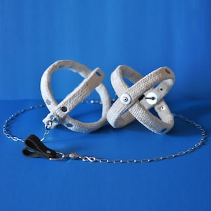 May include: Two beige, woven wrist restraints with silver-tone hardware and a connecting chain. The restraints are connected to a black leather clasp. The background is a solid blue.