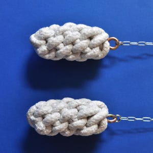 Op de afbeelding: Twee grijze, gevlochten touwdeurgrepen met koperkleurige ringen en zilveren kettingen tegen een blauwe achtergrond. Het touw is strak geweven, waardoor een textuur ontstaat. Deze grepen zijn ontworpen voor functionaliteit en stijl.