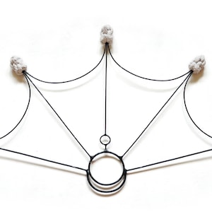 Op de afbeelding: Zwarte metalen wanddecoratie met een geometrisch ontwerp. Het stuk heeft een centraal cirkelvormig element met twee kleinere cirkels en uitstralende lijnen. Vijf grijze, getextureerde, geknoopte elementen bevinden zich aan de uiteinden van de lijnen.