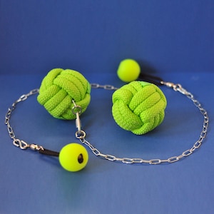 Puede incluir: Un juguete artesanal con bolas anudadas de color verde neón, una cadena plateada y pequeñas bolas amarillas. El juguete está sobre un fondo azul liso. Las bolas anudadas están hechas de cuerda trenzada.