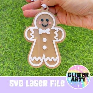 May include: A gingerbread man ornament with white icing and a red bow tie. The ornament is brown and has a white outline. The ornament is held in a hand against a green grass background. The text "SVG Laser File" and "Glitter Arty" are visible in the image.