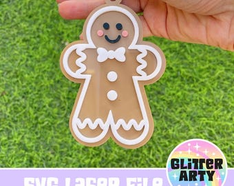 May include: A gingerbread man ornament with white icing and a red bow tie. The ornament is brown and has a white outline. The ornament is held in a hand against a green grass background. The text "SVG Laser File" and "Glitter Arty" are visible in the image.