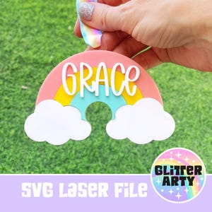 May include: A colorful rainbow-shaped ornament with the name "GRACE" in white script. The rainbow features layers of pink, yellow, and blue, with white cloud accents. The image includes the text "SVG Laser File" and a logo.