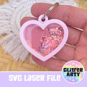 Puede incluir: Un llavero rosa en forma de corazón con un centro transparente lleno de purpurina rosa e iridiscente. El llavero tiene un anillo plateado y el texto "SVG Laser File" está en la parte inferior. La imagen también incluye un logotipo de "Glitter Arty".