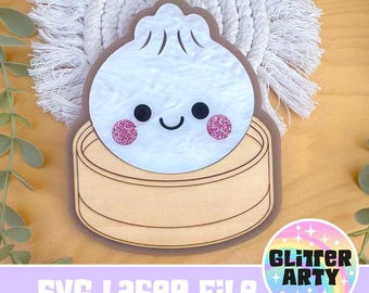 Könnte beinhalten: Ein weißes Acryl-Ornament in Form eines Dumplings mit lächelndem Gesicht und rosa Glitzerwangen, das in einer Basis aus hellem Holz in Bambusdämpfer-Optik ruht. Das Design hat eine kleine Öse zum Aufhängen und einen dunkelbraunen Rand.