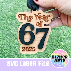 May include: Wooden ornament with the text "The Year of 67 2025" in a retro font. The numbers and text are dark brown against a light wood background. A black ribbon is attached for hanging. The bottom of the image has the text "SVG Laser File".