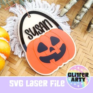 May include: An oval-shaped wooden ornament featuring a black acrylic border and the name SUSAN in black script. A bright orange jack-o'-lantern with black eyes, nose, and mouth is layered over the bottom. It hangs from a red and white string.