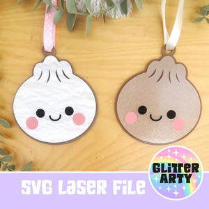 May include: Two hanging ornaments shaped like round dumplings with smiling faces. One is white with a textured surface and a pink polka-dot ribbon, while the other is a metallic bronze colour with a smooth finish and a plain white ribbon.