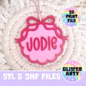May include: A pink 3D printed name tag featuring the name JODIE in raised dark pink lettering. The tag has a scalloped edge, a decorative bow at the top, and a pink ball chain attachment. Text on the image includes 3D PRINT FILE and STL & 3MF FILES.