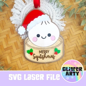 Può includere: Un ornamento decorativo raffigurante un personaggio a forma di gnocco bianco che indossa un cappello da Babbo Natale rosso e bianco, seduto all'interno di un cestello per la cottura a vapore in bambù color legno chiaro con la scritta MERRY Squishmas.