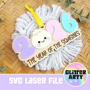 May include: A decorative wooden ornament featuring the number 2026 with a central dumpling character and the text THE YEAR OF THE SQUISHIES. The piece includes a yellow polka dot ribbon for hanging and is layered with pastel purple, pink, and blue.