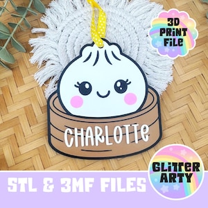 May include: A white dumpling ornament with a smiling face, pink cheeks, and black eyes, sitting in a brown bamboo steamer basket. The name CHARLOTTE is printed in white letters on the front of the basket. A yellow ribbon is attached to the top.
