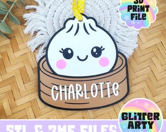 May include: A white dumpling ornament with a smiling face, pink cheeks, and black eyes, sitting in a brown bamboo steamer basket. The name CHARLOTTE is printed in white letters on the front of the basket. A yellow ribbon is attached to the top.