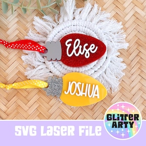 May include: Two Christmas light-shaped ornaments, one red with the name "Elise" and the other yellow with "Joshua". Both have silver glitter accents and are on a white macrame coaster. The background is a woven texture.