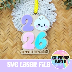 Può includere: Un ornamento in legno con il numero 2026, composto da un 2 viola, uno 0 rosa e un 6 blu. Un raviolo bianco con un volto sorridente è posizionato accanto al 2. Il testo THE YEAR OF THE SQUISHIES è inciso sul fondo, con un nastro giallo.