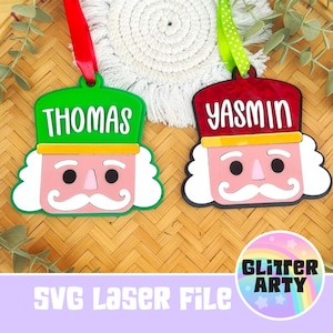 May include: Two nutcracker ornaments with white hair and mustaches. One has a green hat with the name THOMAS and a red ribbon, the other a red hat with the name YASMIN and a green polka-dot ribbon. Both feature a yellow band and pink face.
