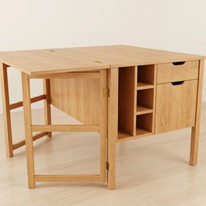 May include: A light-colored wooden drop-leaf table with a cabinet and drawers. The table has a rectangular top with a drop-leaf section, supported by a folding leg. The cabinet features open shelves and two drawers. The table is made of light-colored wood.