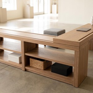 May include: A light brown wooden desk with an L-shaped design. The desk has open shelving, a built-in groove, and a small gray cushion. A black lamp, a white vase, and a laptop are on the desk surface. There are two white boxes, a wicker basket, and a black box on the shelves.
