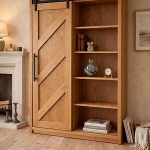 Puede incluir: Librería de roble claro con puerta corredera de granero. La puerta tiene un tirador negro y un diseño diagonal. El lado abierto tiene estantes con libros, un jarrón y un reloj. La librería está sobre un suelo de madera.