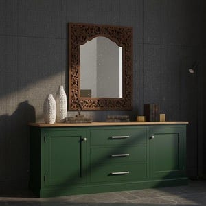 Puede incluir: Un aparador de madera verde oscuro con tres cajones y dos puertas, rematado con una superficie de madera clara. Un espejo de madera tallada cuelga encima, con dos jarrones blancos y libros. La habitación tiene una pared gris oscura.