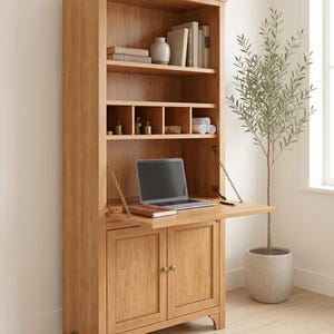 May include: A light oak wooden cabinet with a drop-down desk and shelving. The cabinet features a laptop on the desk, books, and decorative items on the shelves. A small potted tree is to the right.