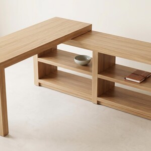 May include: A light-colored wooden desk with a unique L-shaped design. The desk features open shelving for storage and a raised top section. A small bowl and a notebook are visible on the shelves. The desk is made of light-colored wood.