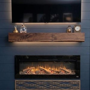 Op de afbeelding: Een donkerblauwe wand met horizontale planken en een gemonteerde televisie boven een natuurlijke houten schoorsteenmantel met een ruwe rand. Op de mantel staan glazen karaffen en een klok. Daaronder een elektrische haard met gloeiende blokken.
