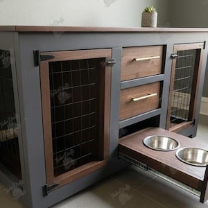 Puede incluir: Un mueble tipo jaula para mascotas en madera gris oscuro con tapa de madera marrón, dos puertas de malla metálica, dos cajones con tiradores dorados y un cajón extraíble con dos cuencos de acero inoxidable.